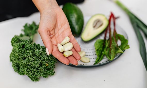 Close-up of a hand holding garlic cloves over a plate with fresh vegetables, including kale and avocado.
