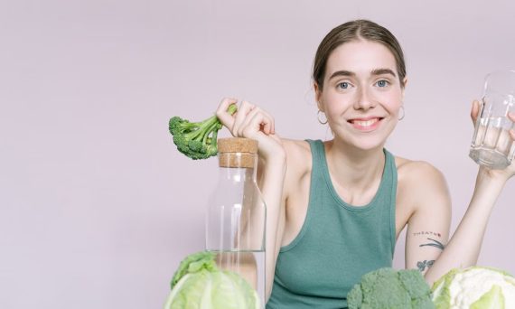 Woman in tank top smiling with broccoli, cabbage, and glass of water on table.